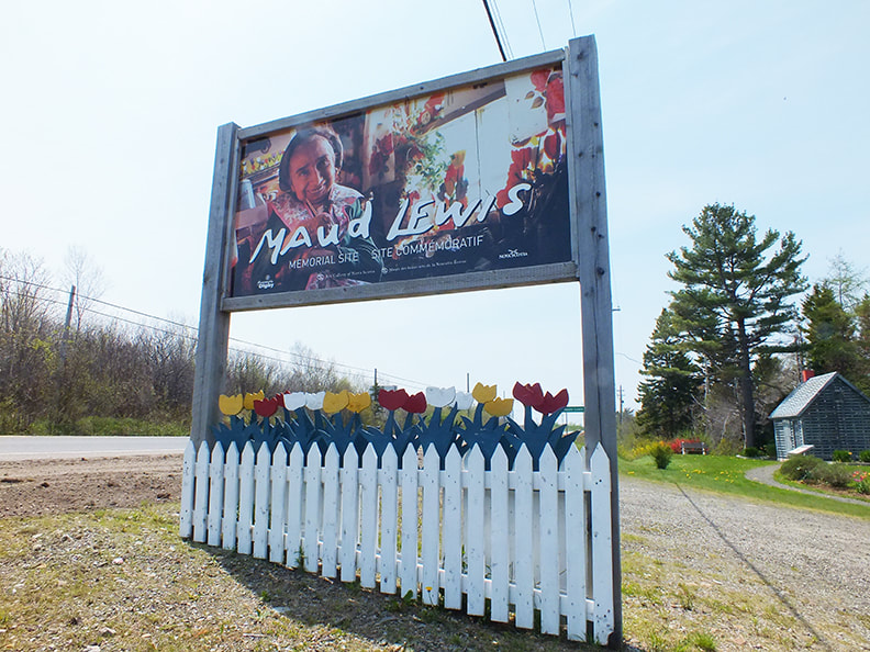 A roadside sign advertising the Maud Lewis Memorial, which has a picture of the artist and painted wooden tulips at the base, with the grey memorial house in the background.Picture