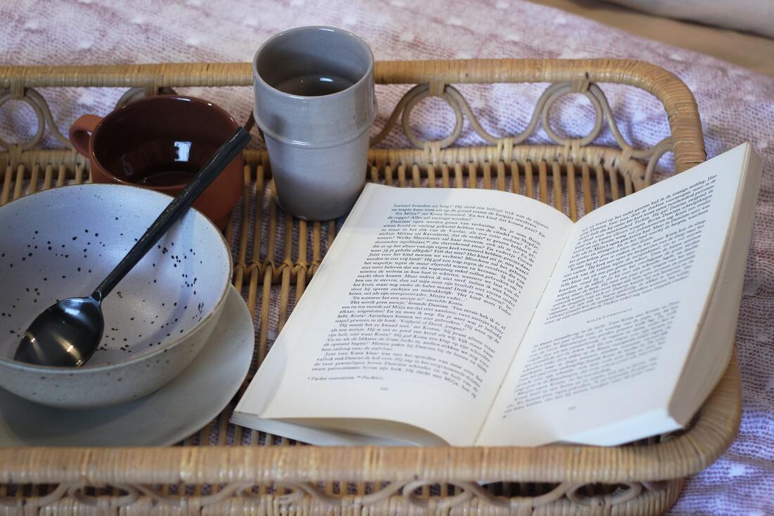 Wicker tray with an open book, blue speckled bowl, and mugs. Picture