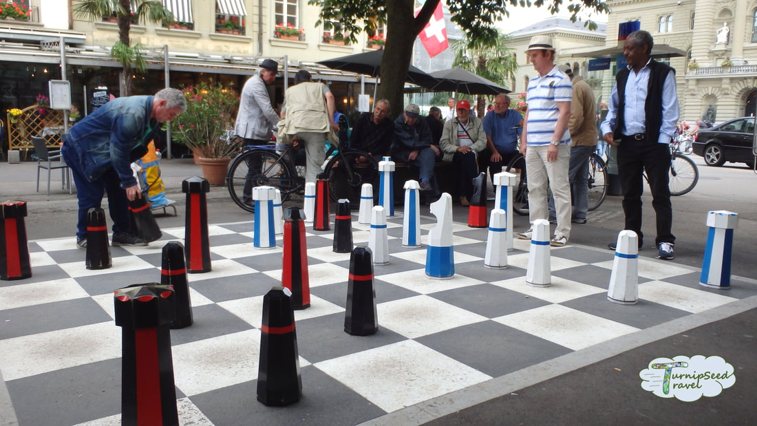 Men play a game of giant chess on a huge board in a public square.