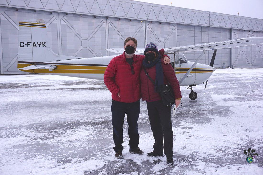 Vanessa and Ryan, both wearing red jackets and black face masks, get ready to board the small plane on a snowy day 