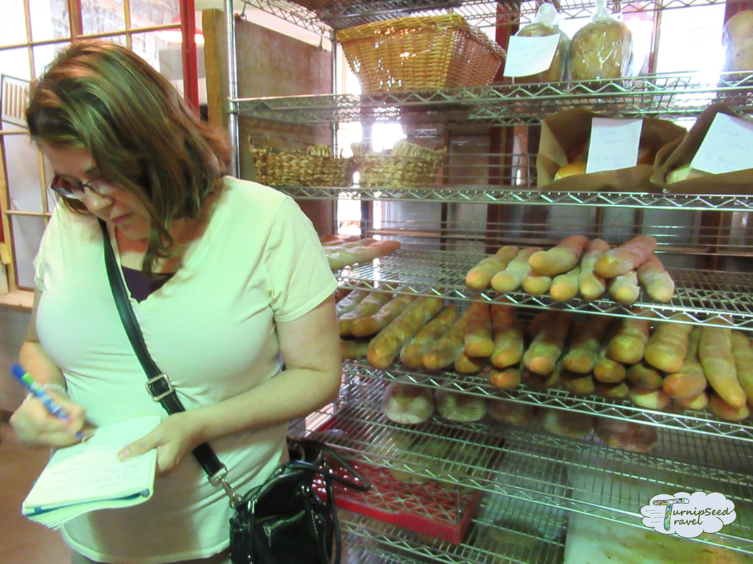 Vanessa takes notes in a bakery while wearing a yellow shirt with a shelf of bread loaves behind her Picture
