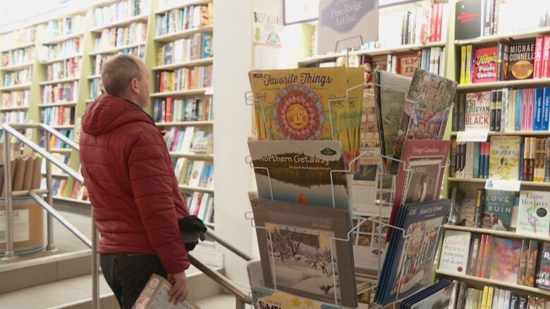 Man in a red winter coat looks at a long wall of books and there is a rack with calendars in the foreground.