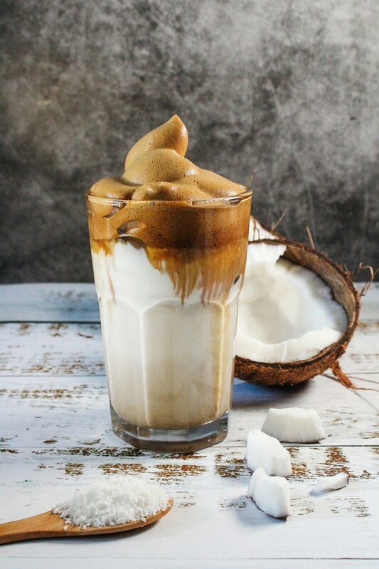 Dalgona coffee in a tall clear glass with half a fresh coconut cracked in half and lying beside the glass.Picture