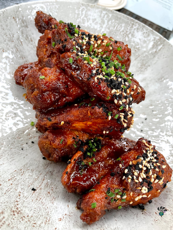 Chicken wings in a thick red sauce and sprinkled with sesame seeds, black sesame seeds, and herbs sit on a silver serving platter