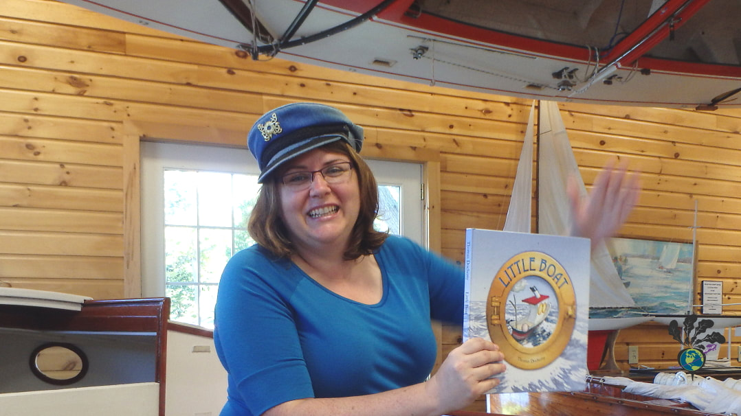 Vanessa sits in a boat and holds up a kid's book while wearing a blue shirt and a blue sailor's cap at the Lawson Center Boat Museum Picture