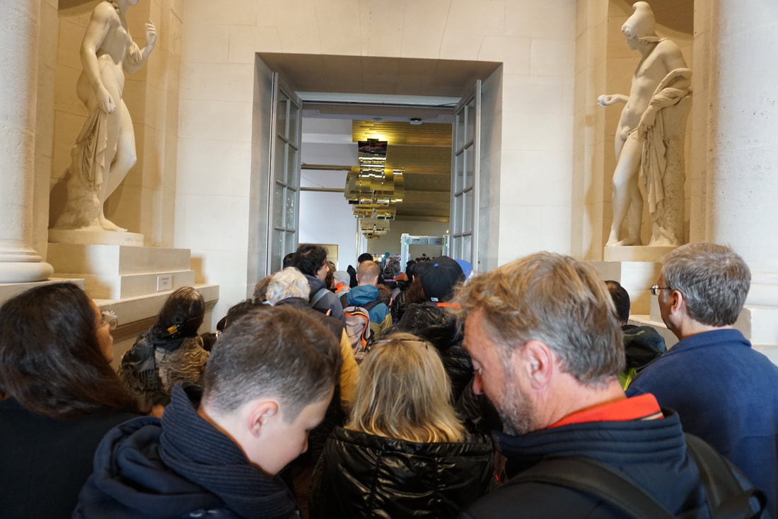 Overview of crowds in a corridor waiting to go through a security scanner, with marble statues on both sides of the wall.