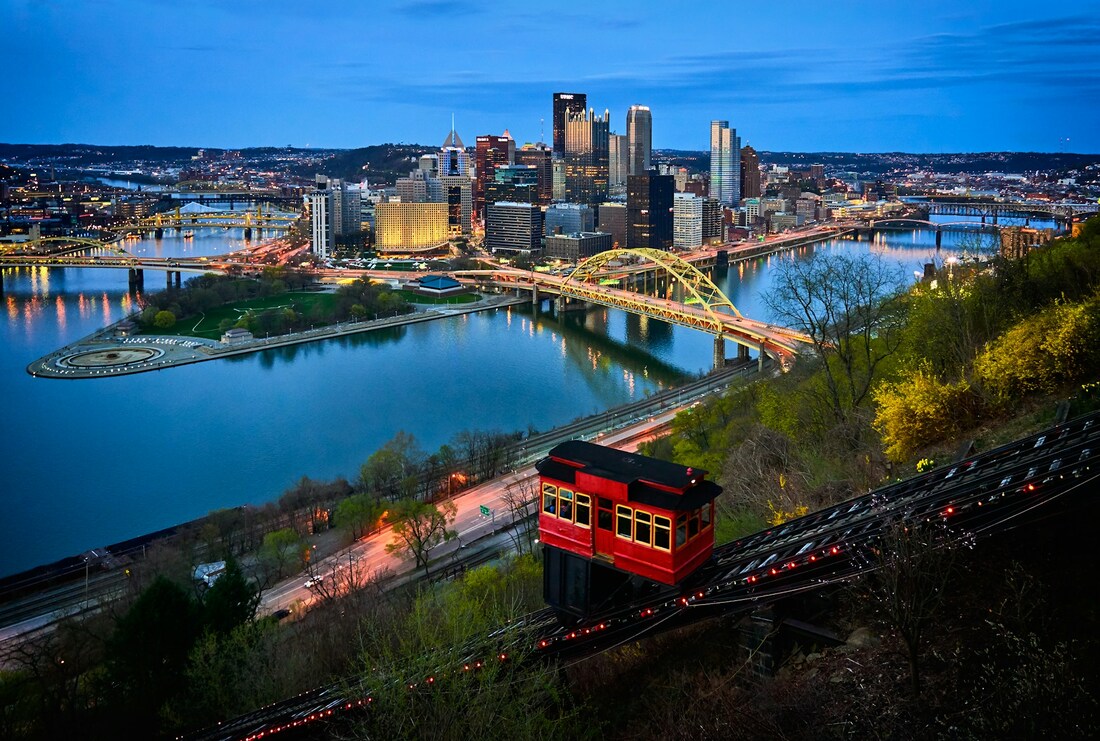 A red funicular climbs up a hill with the Pittsburgh skyline and bridges in the background at dusk with a dark blue sky
