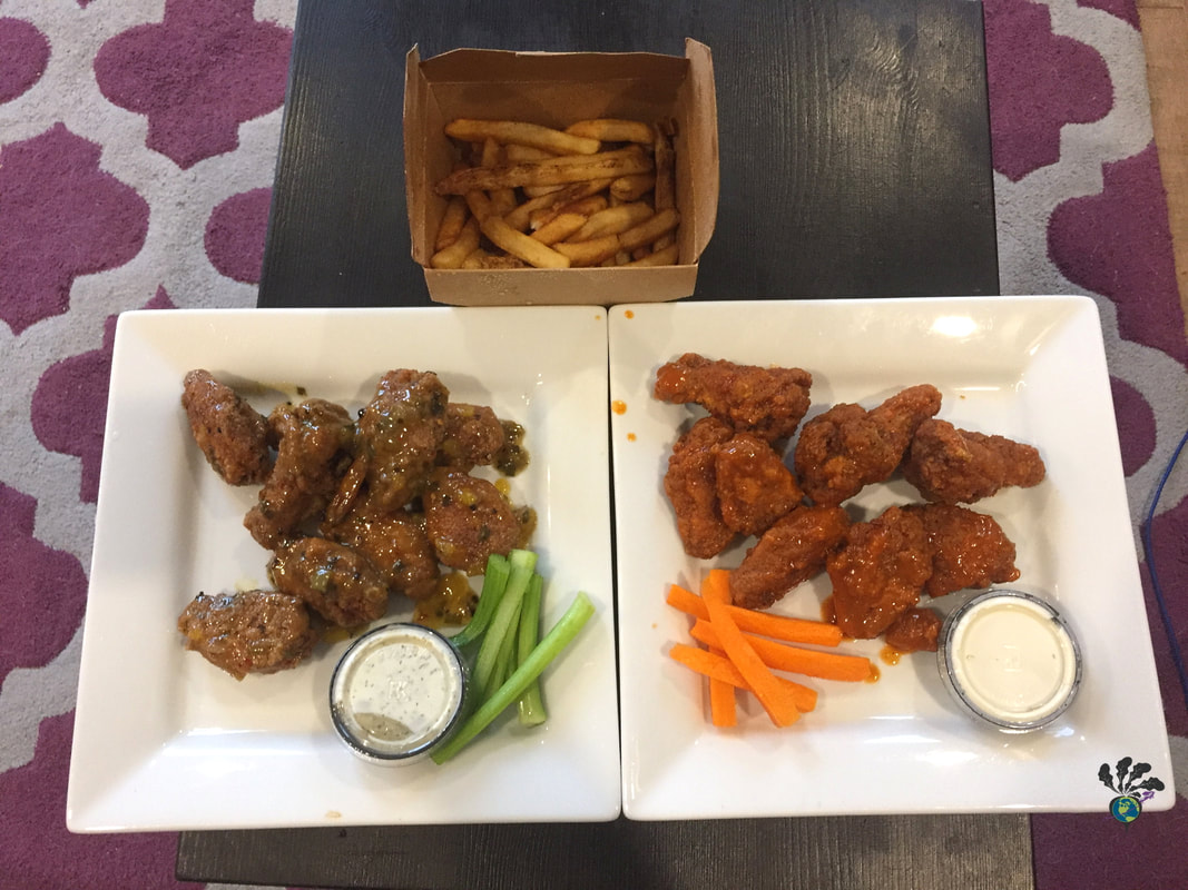 Two square white plates sit on a black coffee table, each containing wings, a container of dip, and some veggies sticks. A take out box of fries is beside them.Picture