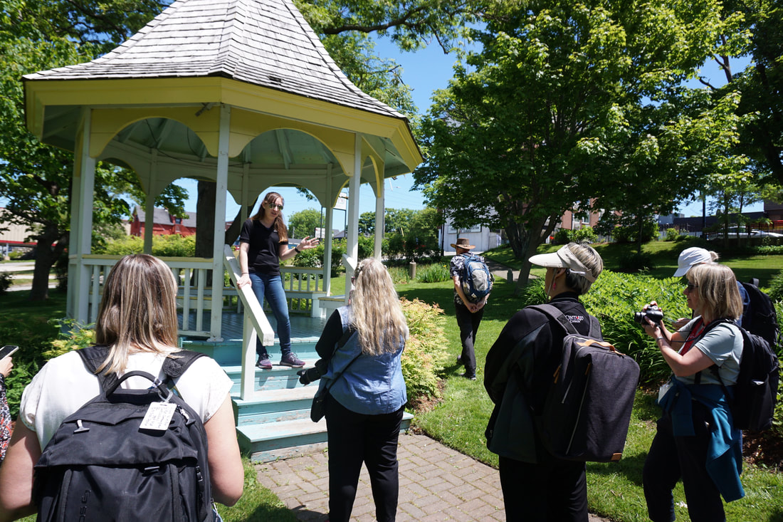 Tourguide stands in a park pavilion, wearing a black shirt and sun glasses, while visitors stand around her in a semi-circle, some taking photos.