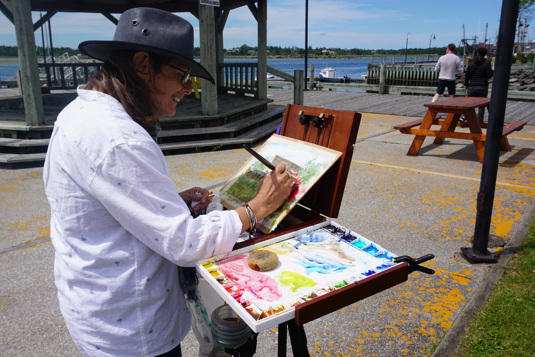Woman in a white shirt and blue hats paints watercolours while standing at an easel by Yarmouth's harbour, with a wooden pavilion, boats, and the water in the background.
